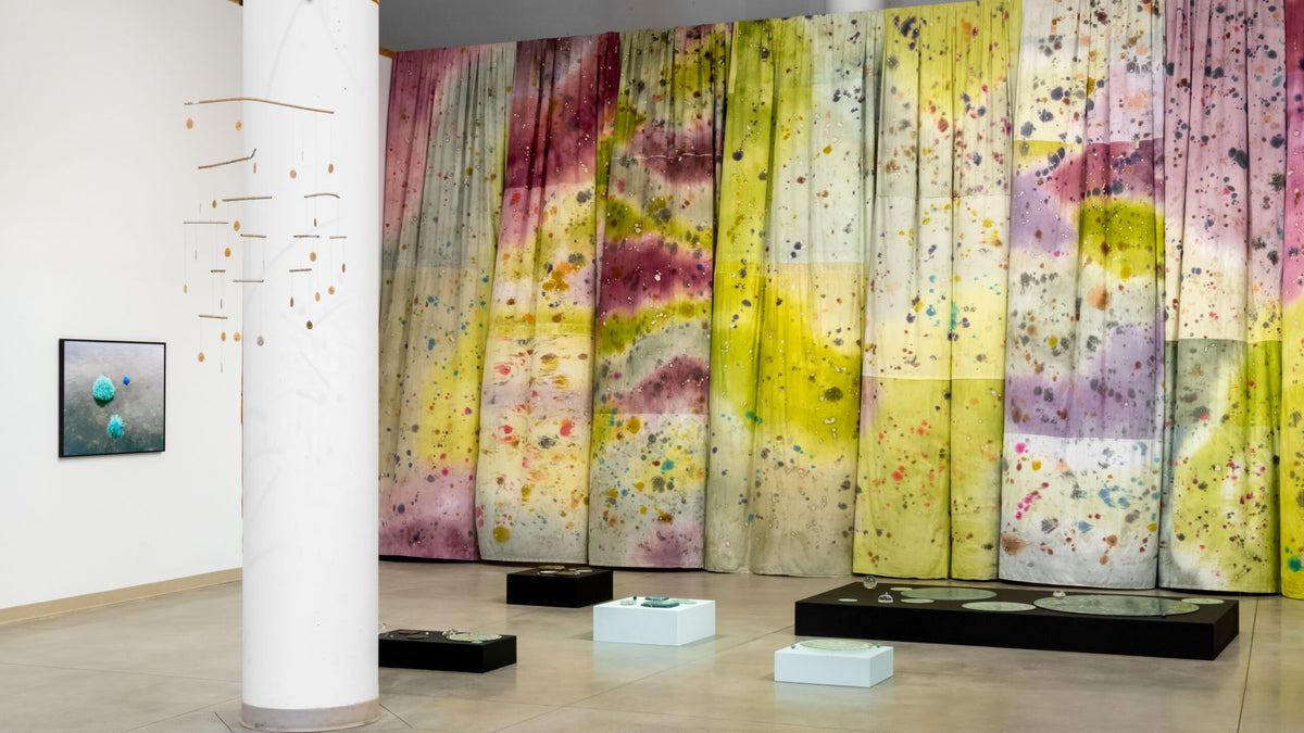 Floor to ceiling fabric panels dyed yellow, pink and purple hang behind low plinths of glass pieces and a hanging mobile