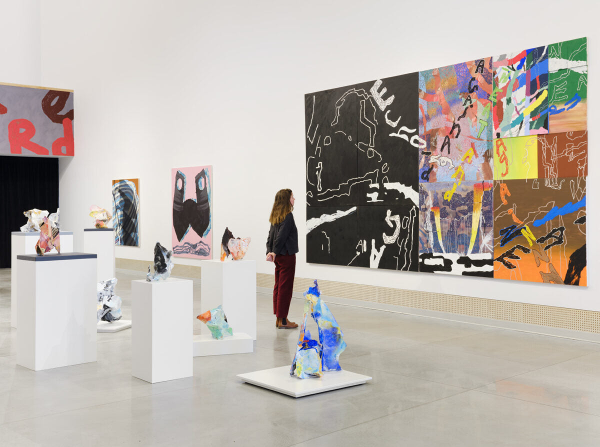 A visitor looks at a large painting of patchwork panels and lettering, with small painted sculptures on white plinths