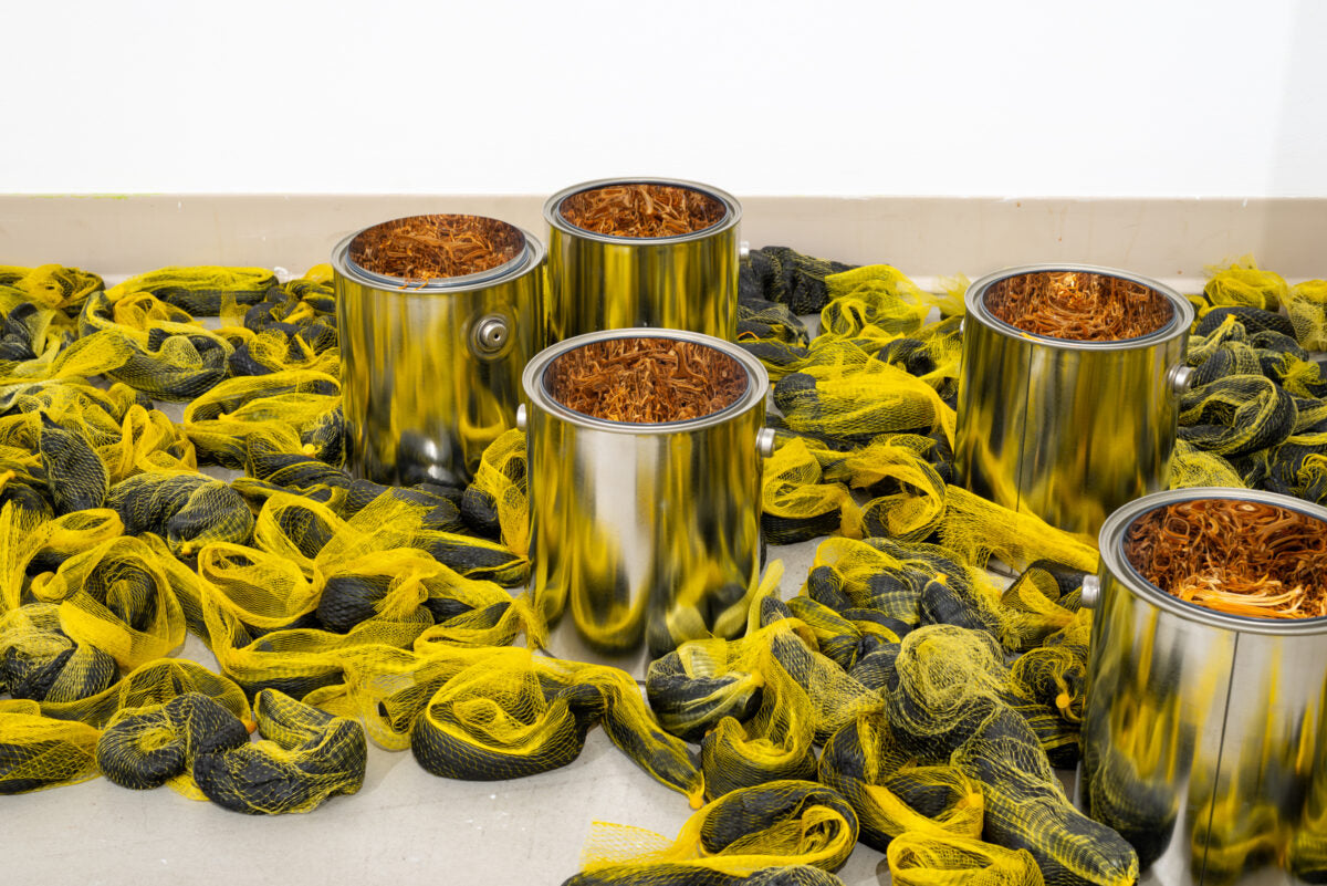 An artwork by Lotus Kang featuring shiny metal cans filled with copper-coloured strands, standing among bundles of yellow and black netting on the floor