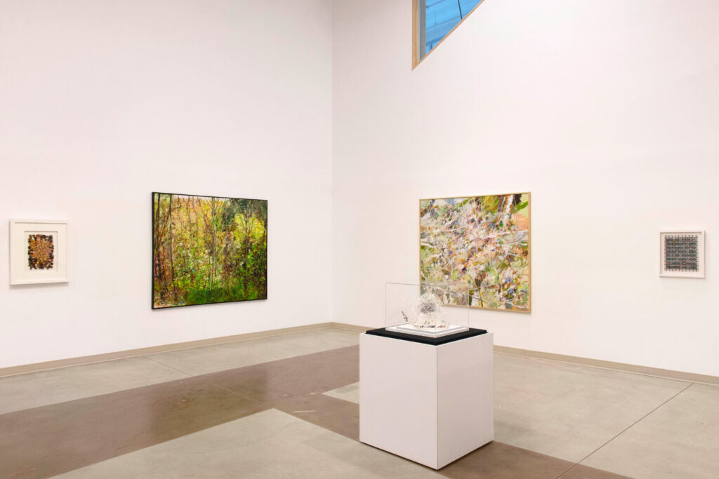 Two large landscape paintings and two small framed works hang around a small sculpture in a clear case on a plinth