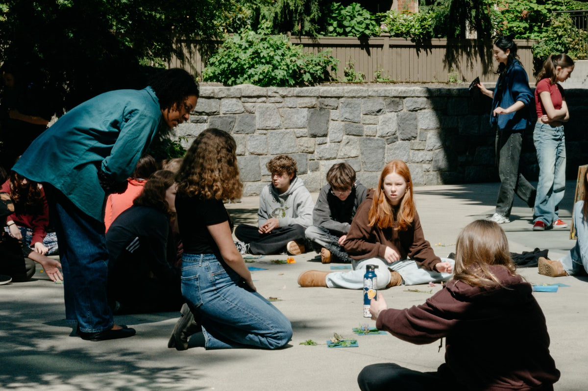 Artist Rebecca Bair talks with students sitting on the ground in a courtyard during her 2026 Artist in Residence workshop