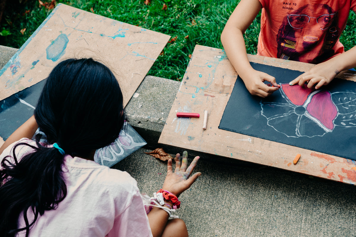 Two children draw with chalk pastels on black paper outdoors, one colouring a large flower