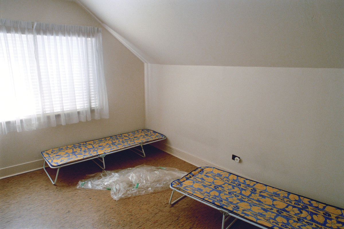 Colour photograph of a bare attic room with two folding cots in yellow and blue floral fabric and a sheet of plastic on the floor