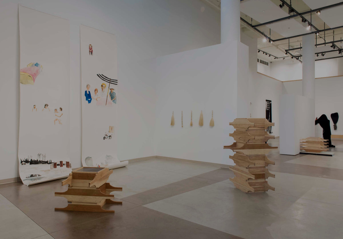 Two long paper scrolls painted with small scenes of people hang to the floor beside stacked wooden sculptures and five small brooms on the wall