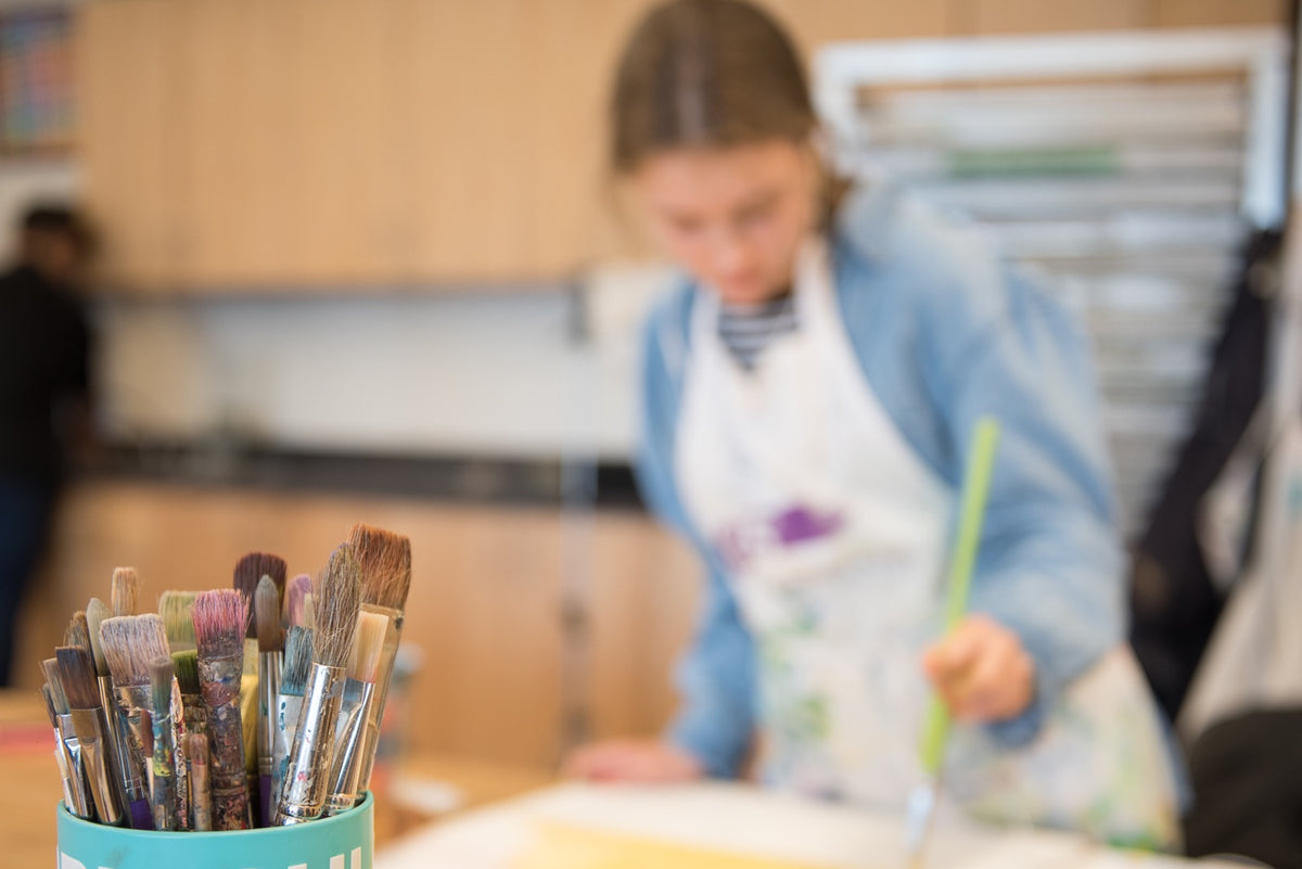 A student in a paint-spattered apron paints at a studio table, out of focus behind a cup of well-used paintbrushes