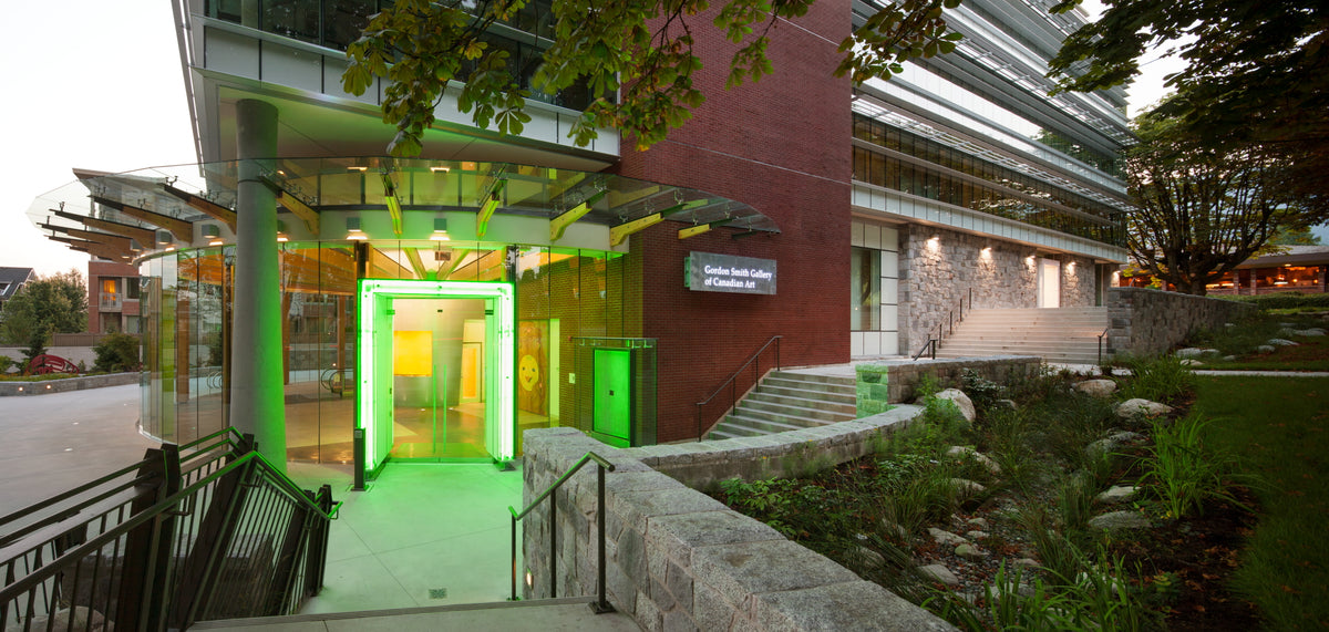 The gallery entrance at dusk, a doorway lit bright green under a glass canopy, beside brick and stone walls