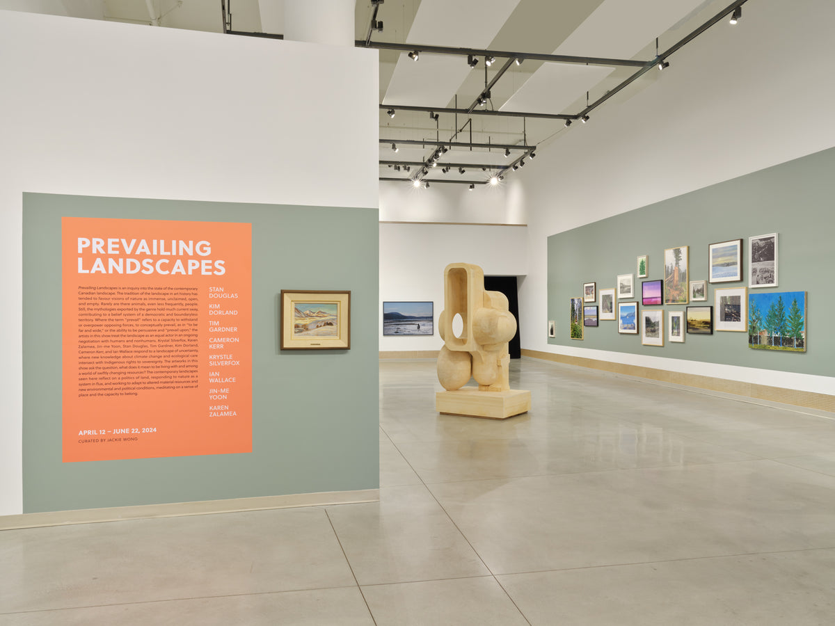 An orange introduction panel on a grey-green wall, a pale wood sculpture, and a wall of closely hung landscape pictures