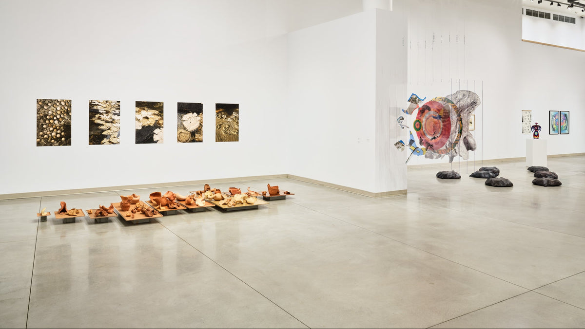 Five photographs hang above trays of clay forms on the floor, with a hanging collage and carved dark forms beyond