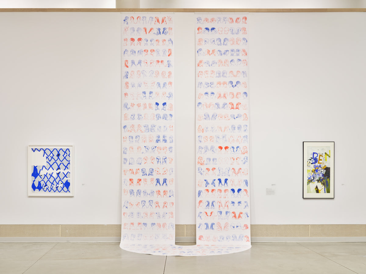 Two long paper scrolls printed with rows of small red and blue heads hang to the floor between two framed works