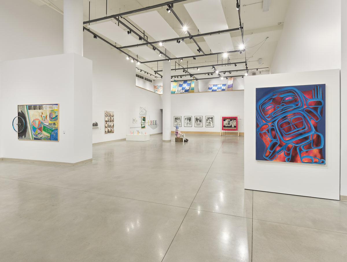 Paintings on freestanding walls, one a blue formline figure on red, with quilts hung high and framed works beyond