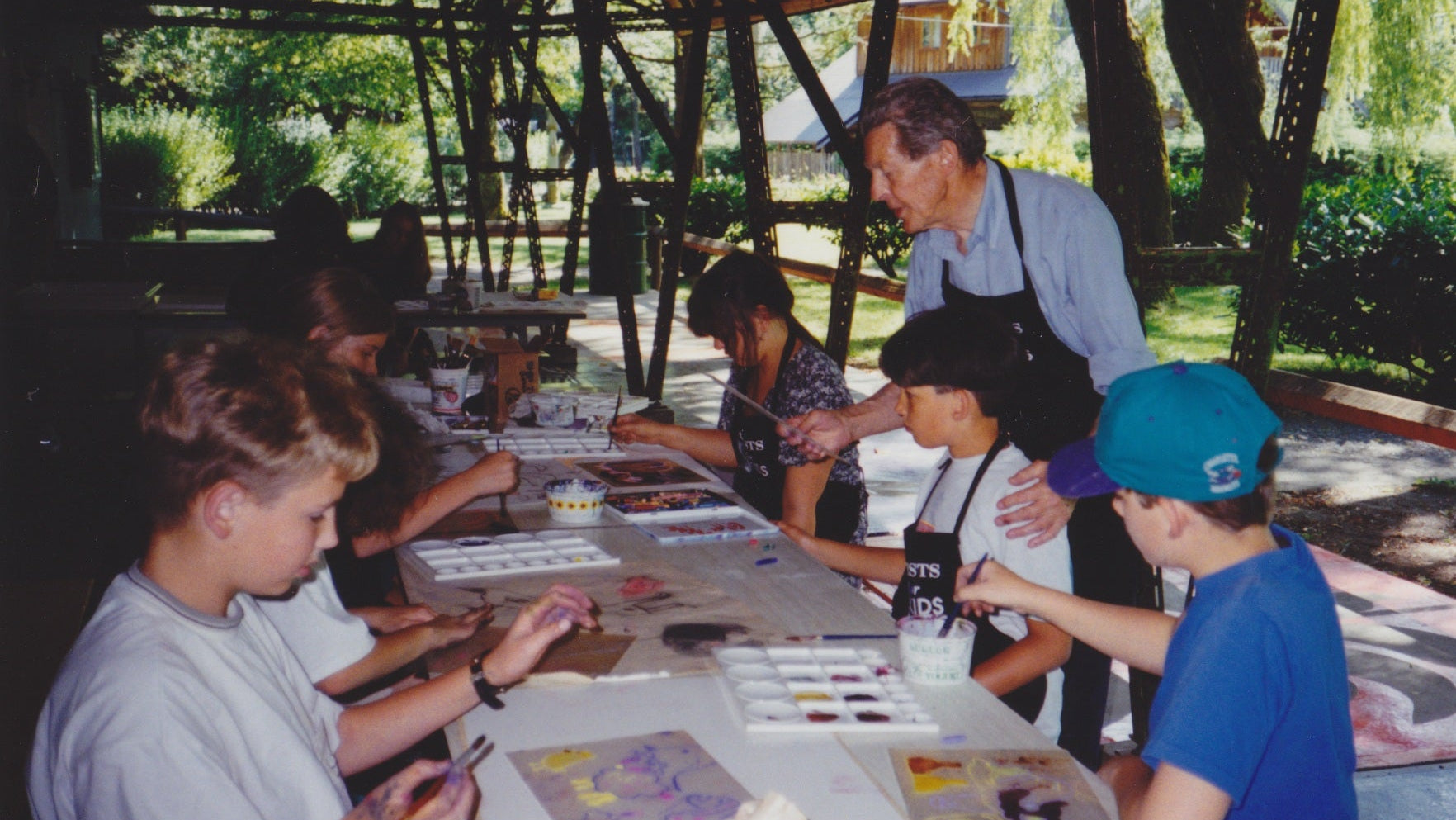 At Paradise Valley Summer Camp in 1994, a group of children painting at an outdoor table with Gordon Smith in a light blue shirt and black apron standing and guiding the art activity.