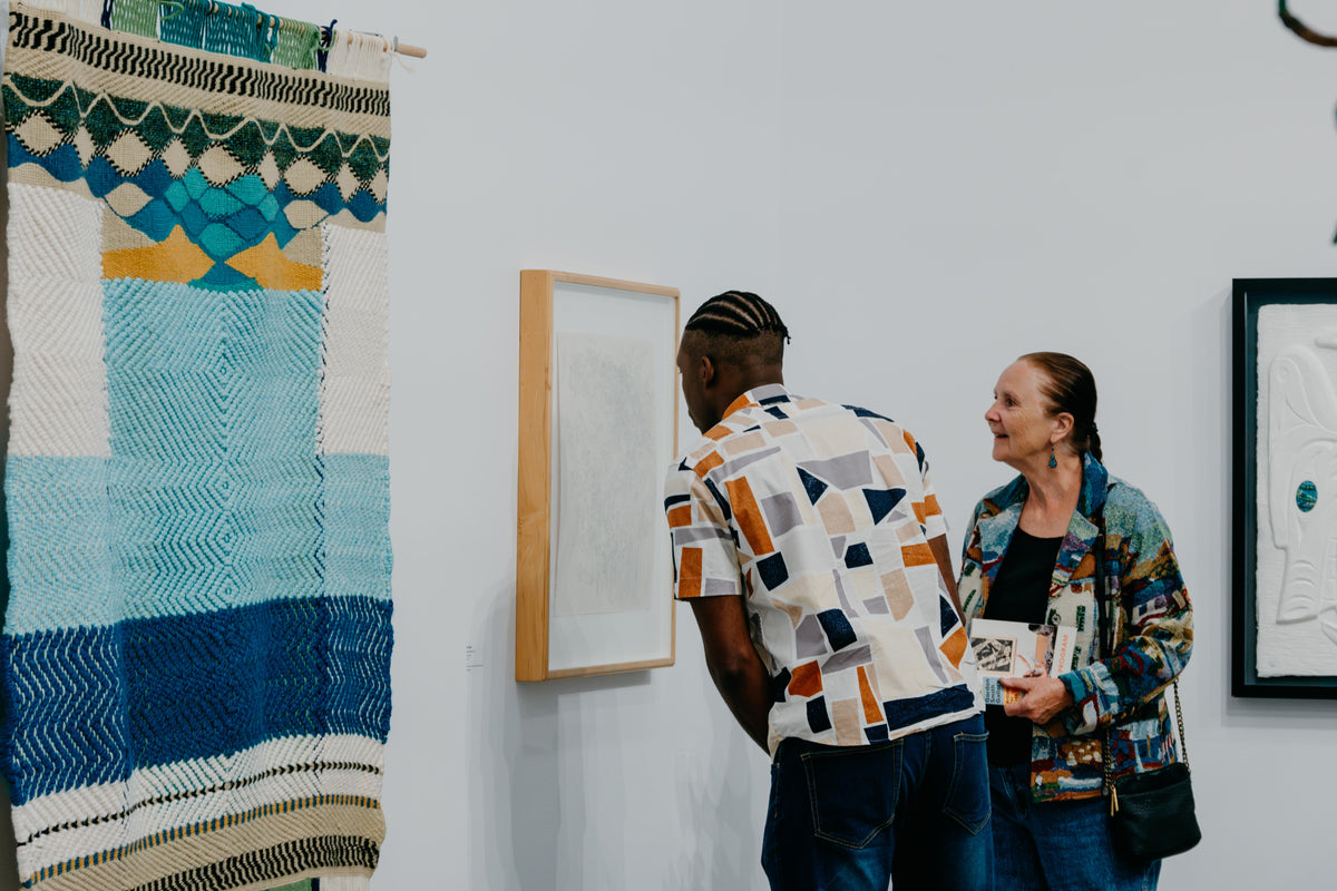 Two visitors look closely at a framed drawing, beside a large woven hanging in blue, green and white