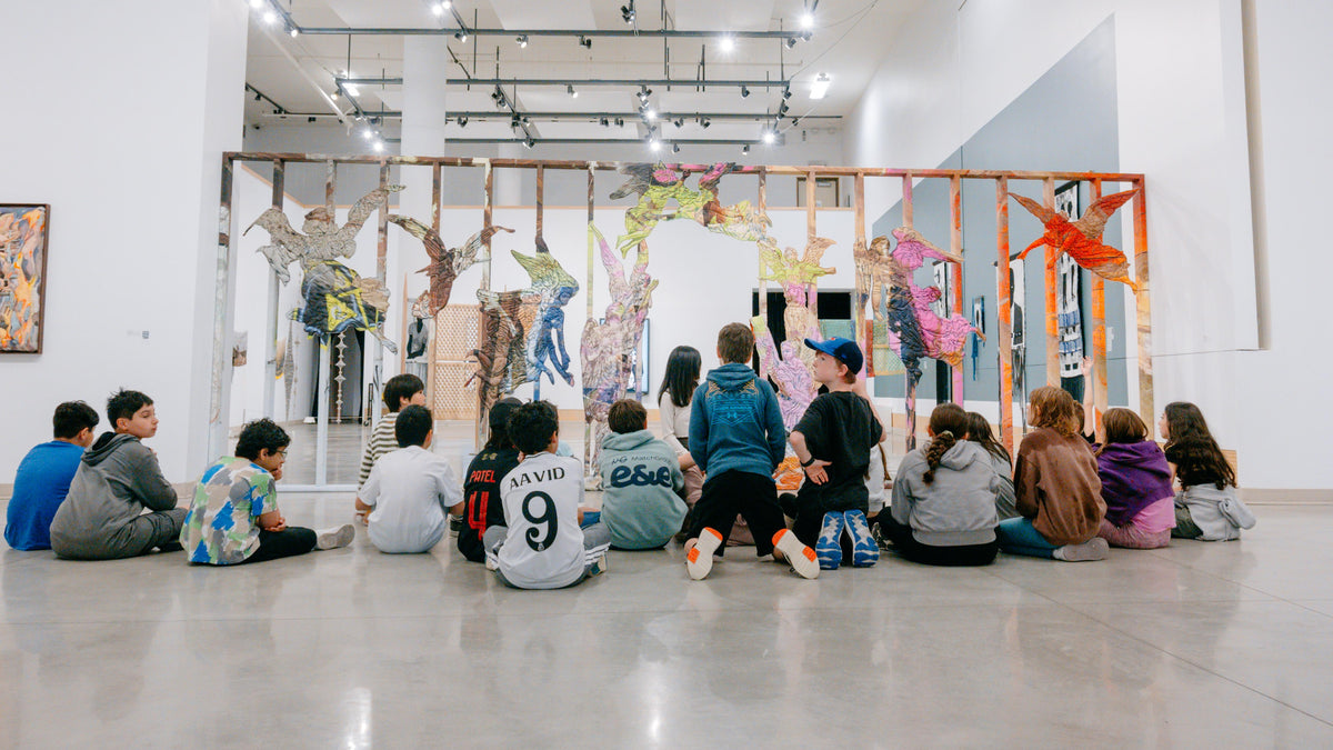 A group of children sit on the gallery floor facing an art educator in front of a large wooden frame covered in colourful cut-out figures by artist Maya Beaudry