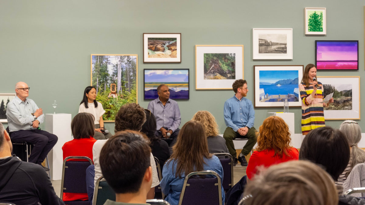 A speaker with a microphone introduces four seated panelists to an audience, in front of a wall of landscape pictures