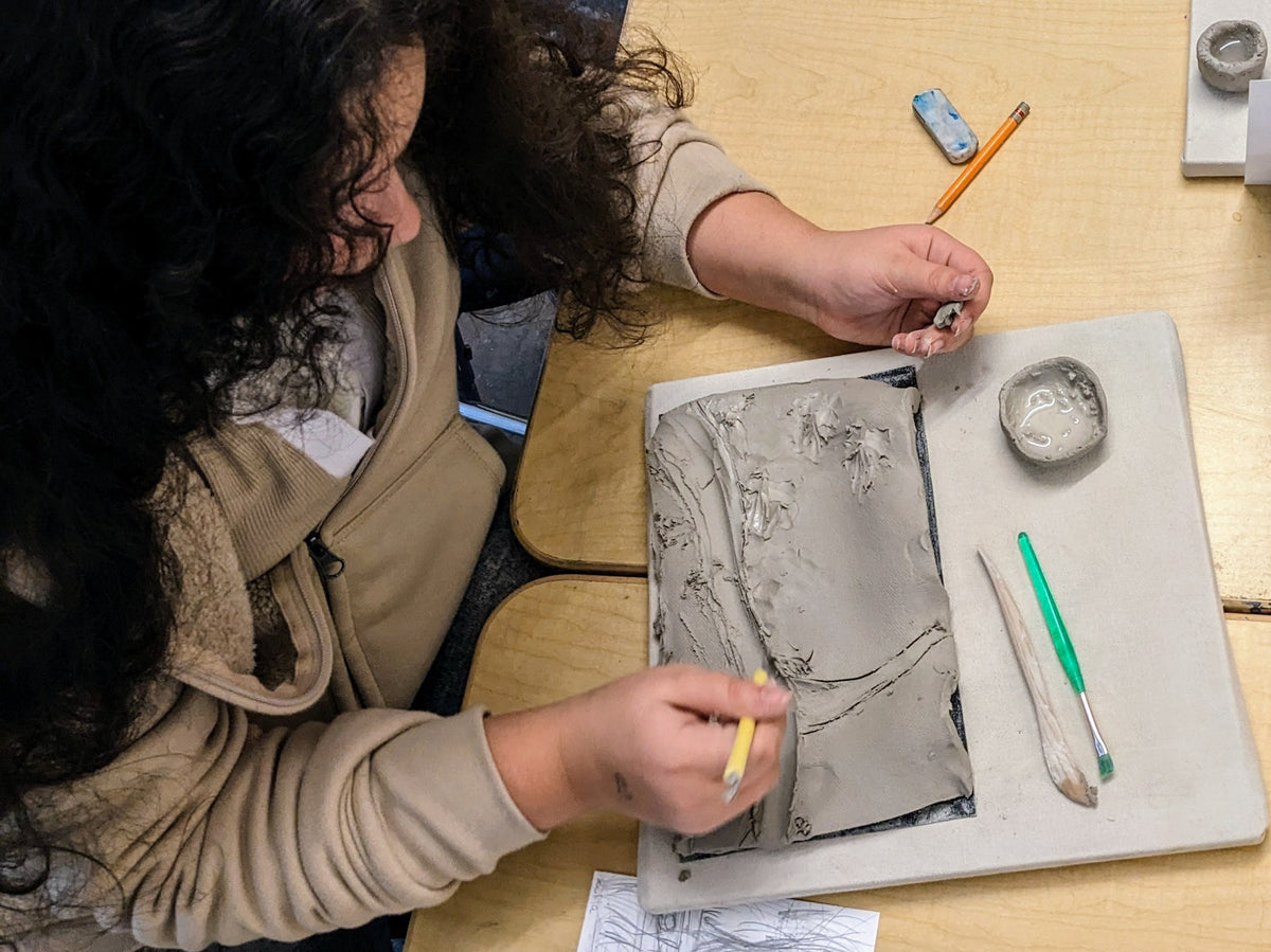 A student pressing a tool into a clay tile at a classroom desk
