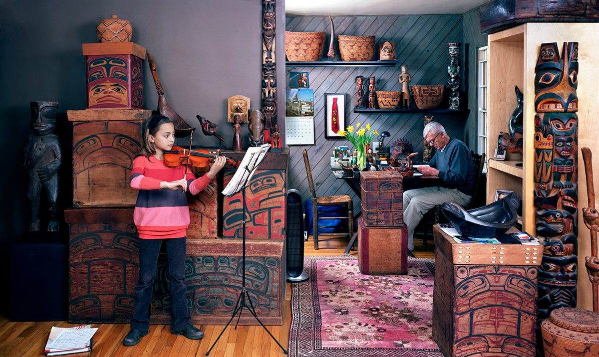 Christos Dikeakos, The Music Lesson, 2013. A little girl practicing violin in a room with bentwood boxes. A man sits in a neighbouring room surrounded by first nations artifacts.