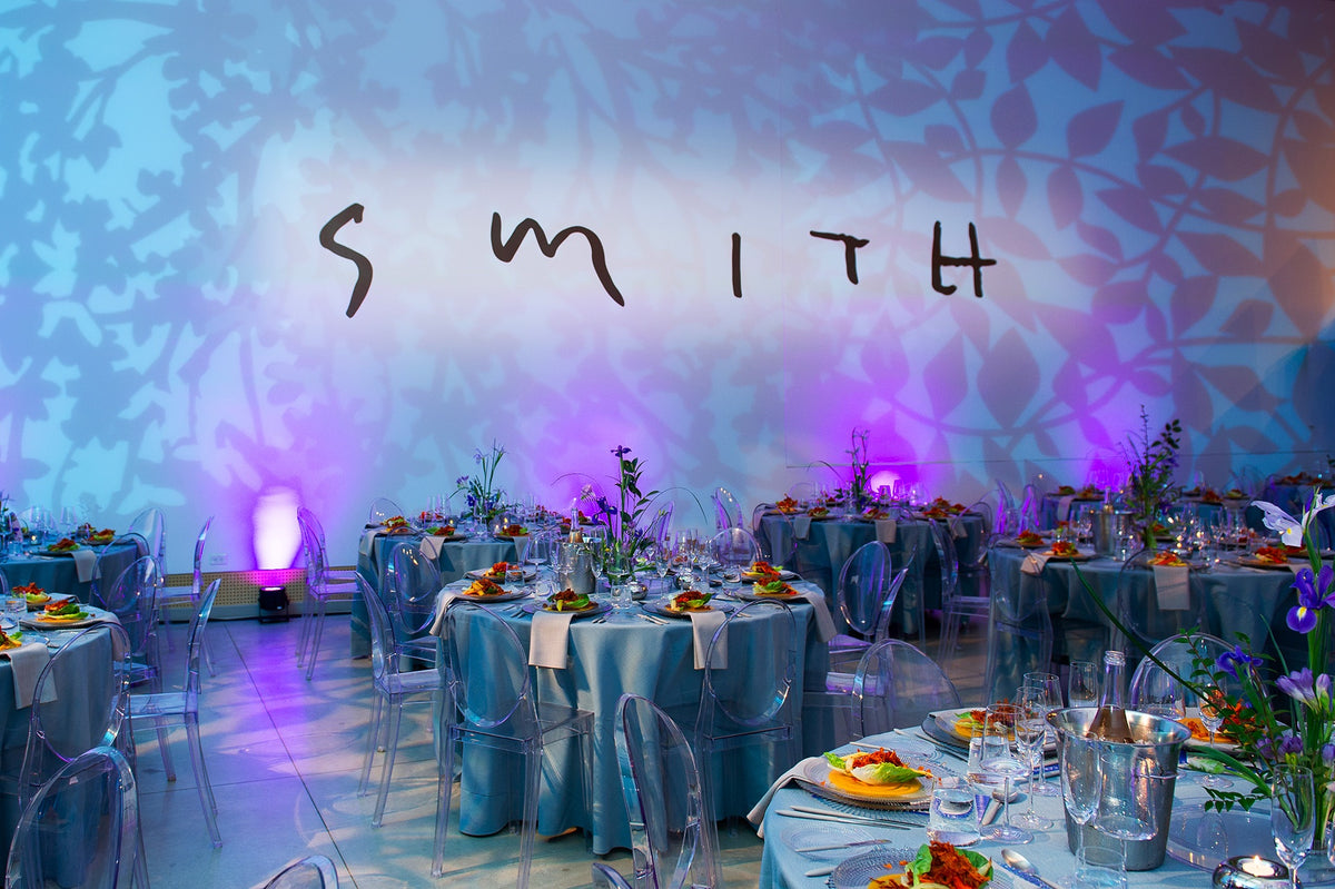 Round tables set for dinner with blue cloths, clear chairs and irises, below the word Smith on a wall lit with leafy shadows
