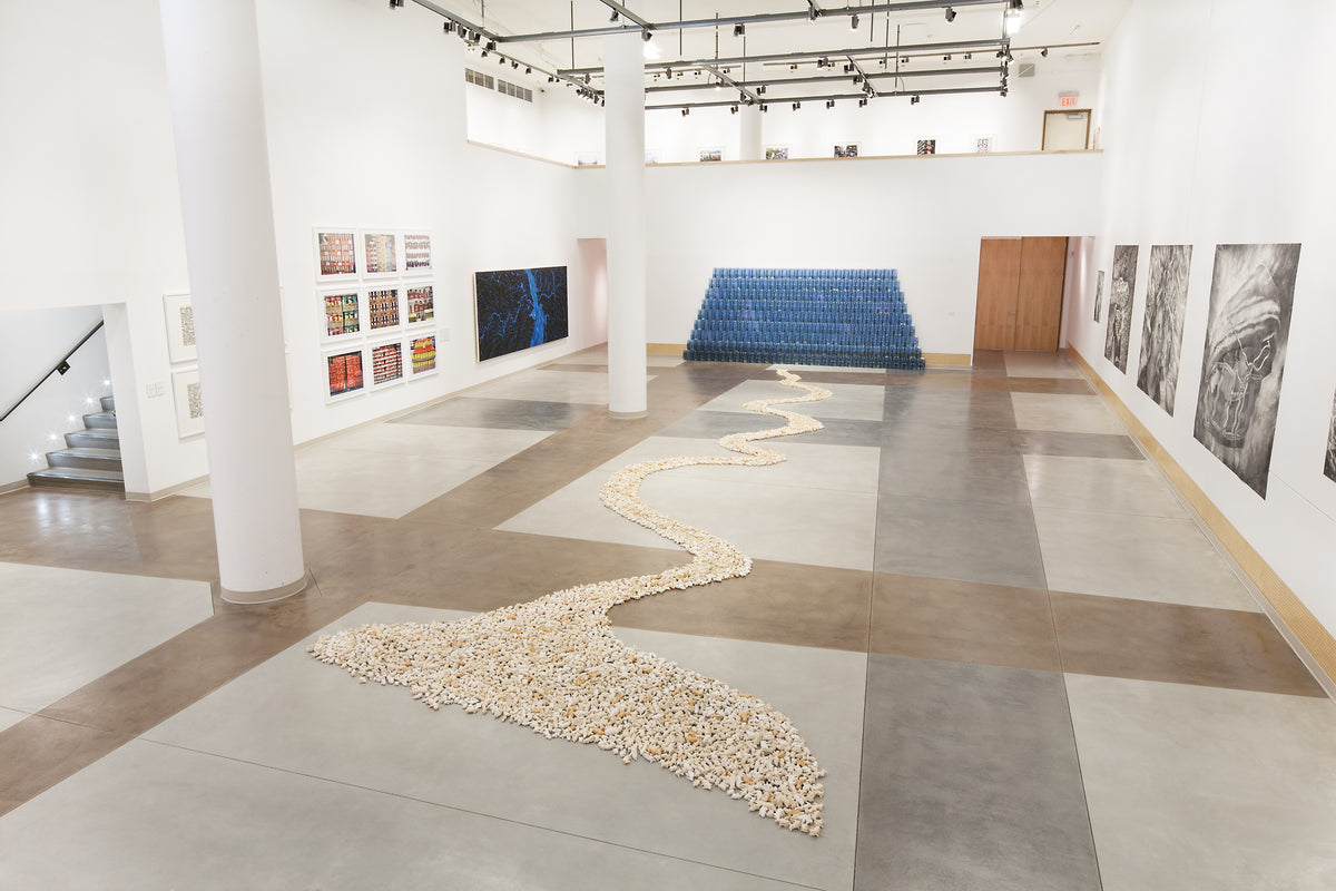 A winding river of small white pieces crosses the gallery floor to a stepped wall of blue plastic jugs, with photographs and drawings on the walls