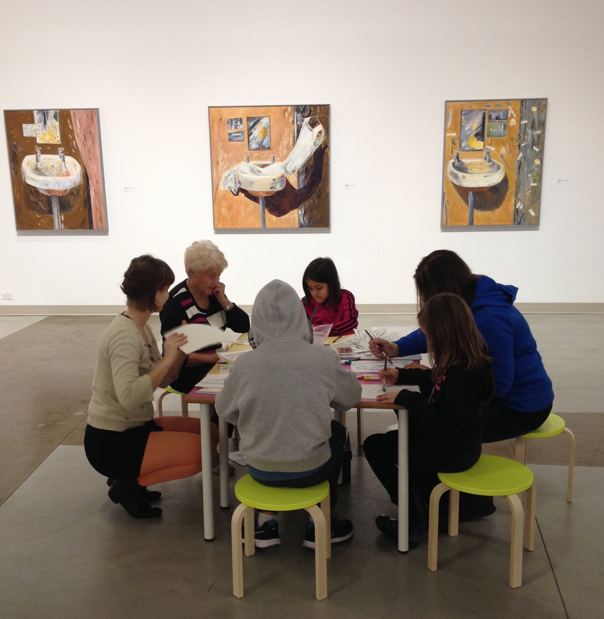 Children and adults draw together at a table with bright green stools, below three paintings of washbasins on warm orange walls