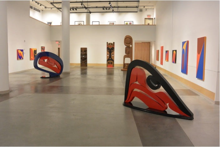 Large red, black and blue sculptures of curved formline shapes stand among carved wooden poles and bright abstract paintings