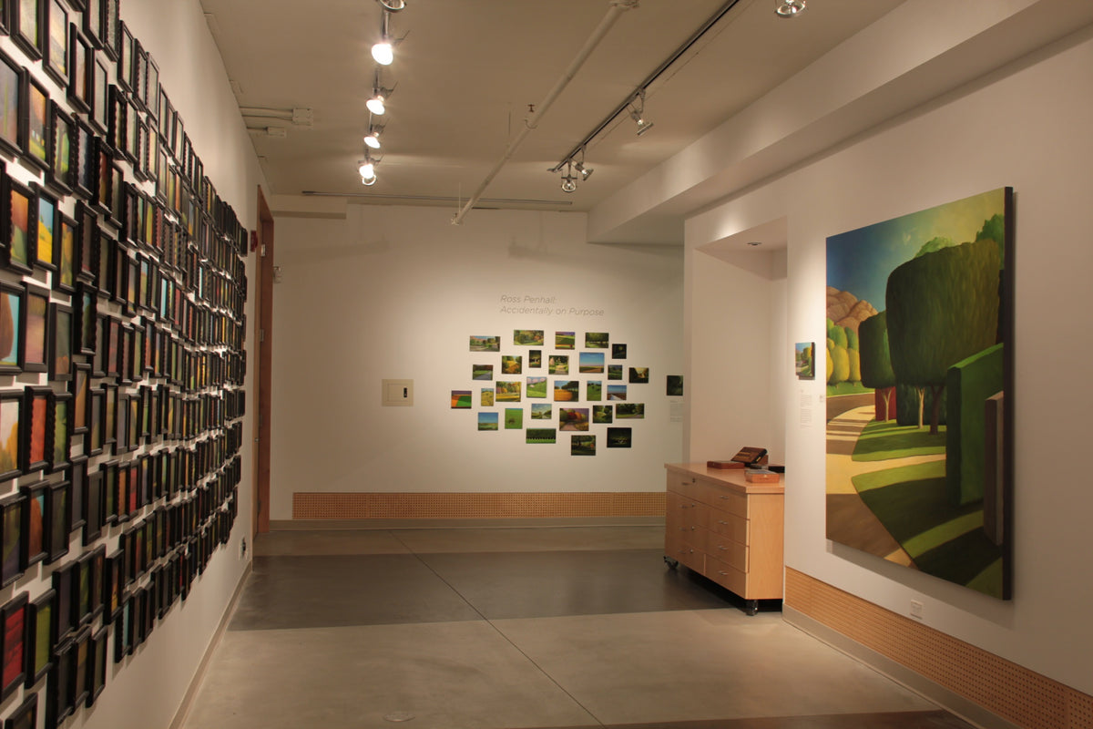 A wall packed with small framed landscape paintings faces a large painting of trimmed green trees and a cluster of small landscapes