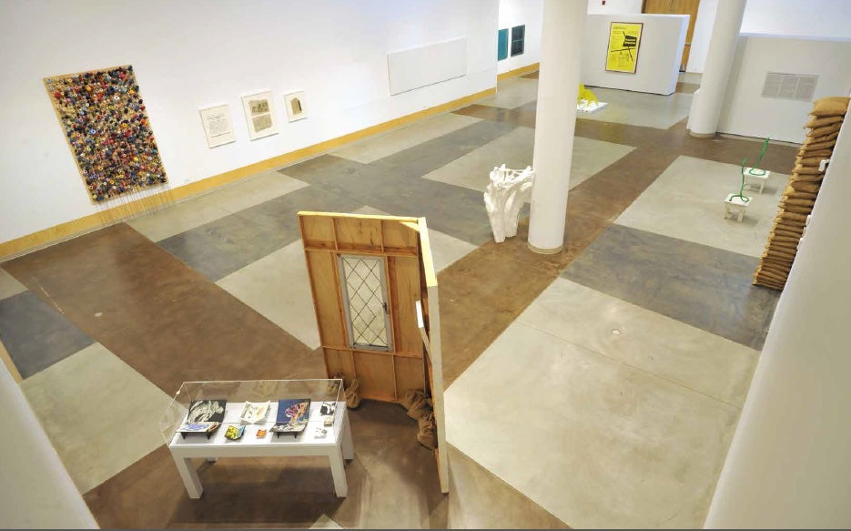 Seen from above, a gallery with a wooden wall section holding a leaded window, a glass case of small objects, a stack of sandbags and wall works