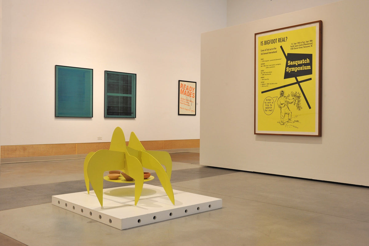 A yellow metal sculpture holding clay bowls stands on a white plinth, near a framed yellow poster about Bigfoot and two teal prints