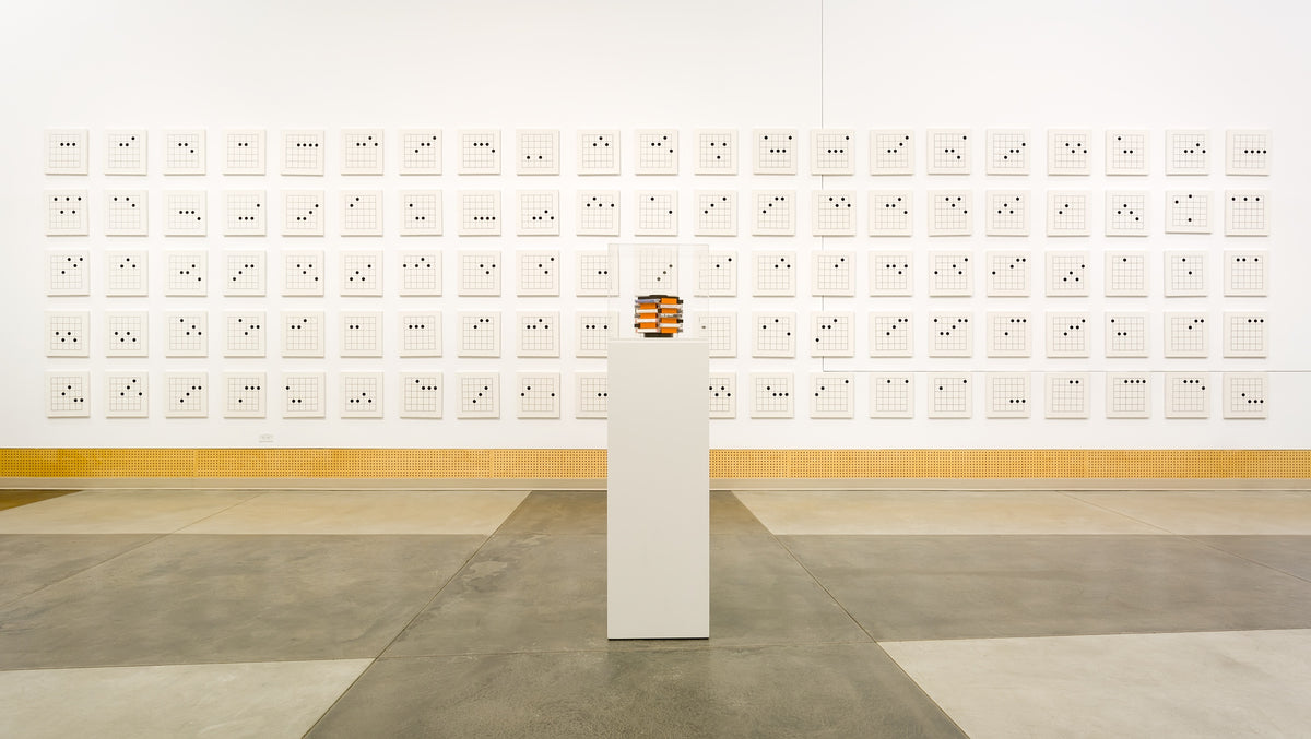 A long wall of small square grid drawings, each marked with a few black dots, behind a plinth holding a small orange object in a clear case