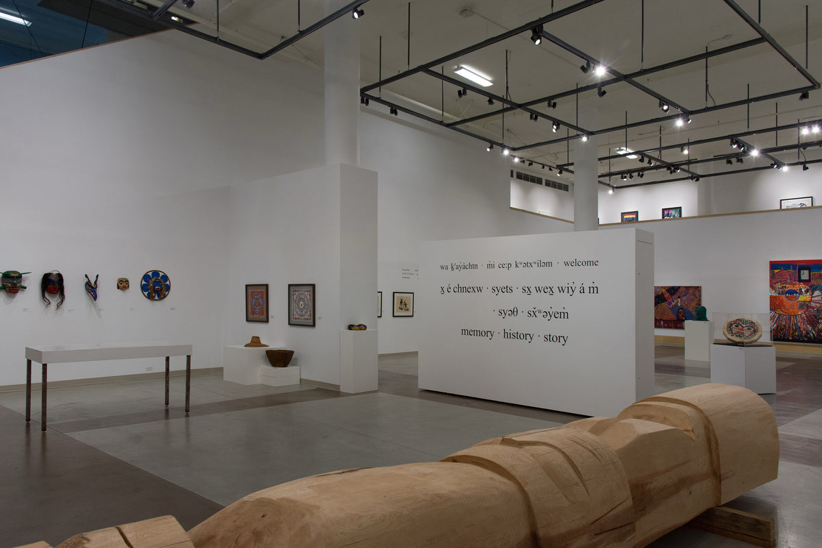 A carved cedar log lies in front of a wall of welcome words in Indigenous languages and English, with masks, prints and baskets beyond
