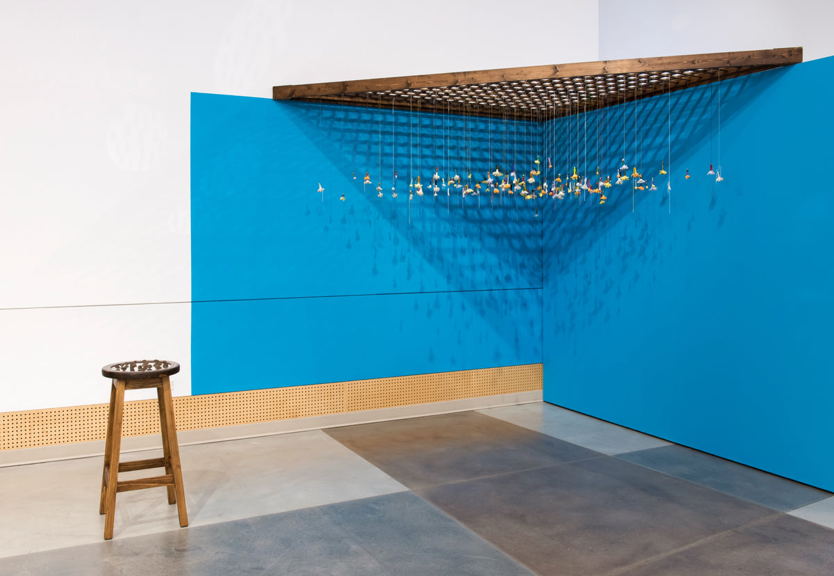 Dozens of small coloured objects hang on threads from a wooden grid in a corner painted bright blue, with a wooden stool nearby
