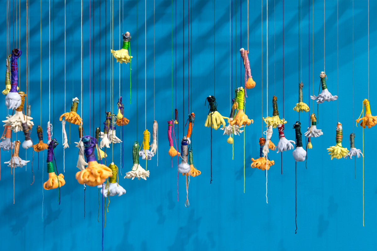 Close view of small hand and flower shapes in yellow, orange and white, wrapped in coloured thread and hanging on strings against a blue wall