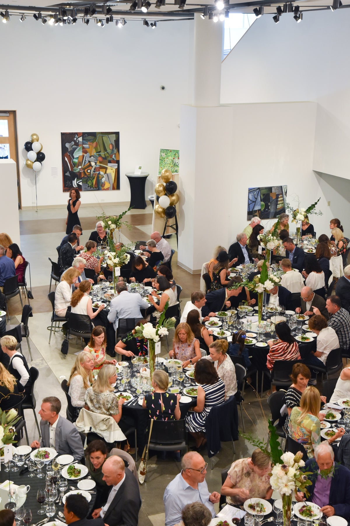 Seen from above, guests dine at round tables with tall white flower centrepieces in a gallery hung with paintings and balloons
