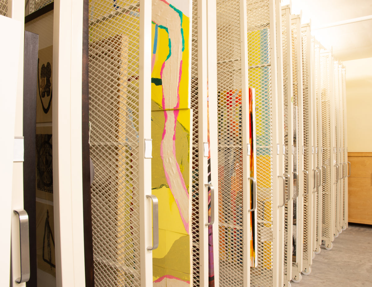 Sliding mesh storage racks in the art vault, with a bright yellow and pink painting by artist Russna Kaur on the nearest rack